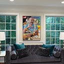 A framed colorful Jimmy Buffet collage portrait of a baseball player hangs on a light gray wall above a gray tufted sofa in a styled living room with two windows, lamps, and pillows.