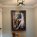 A large framed wall art print depicting an angel with white wings and dark hair, hanging on a gray wall in a home hallway above a wooden banister.