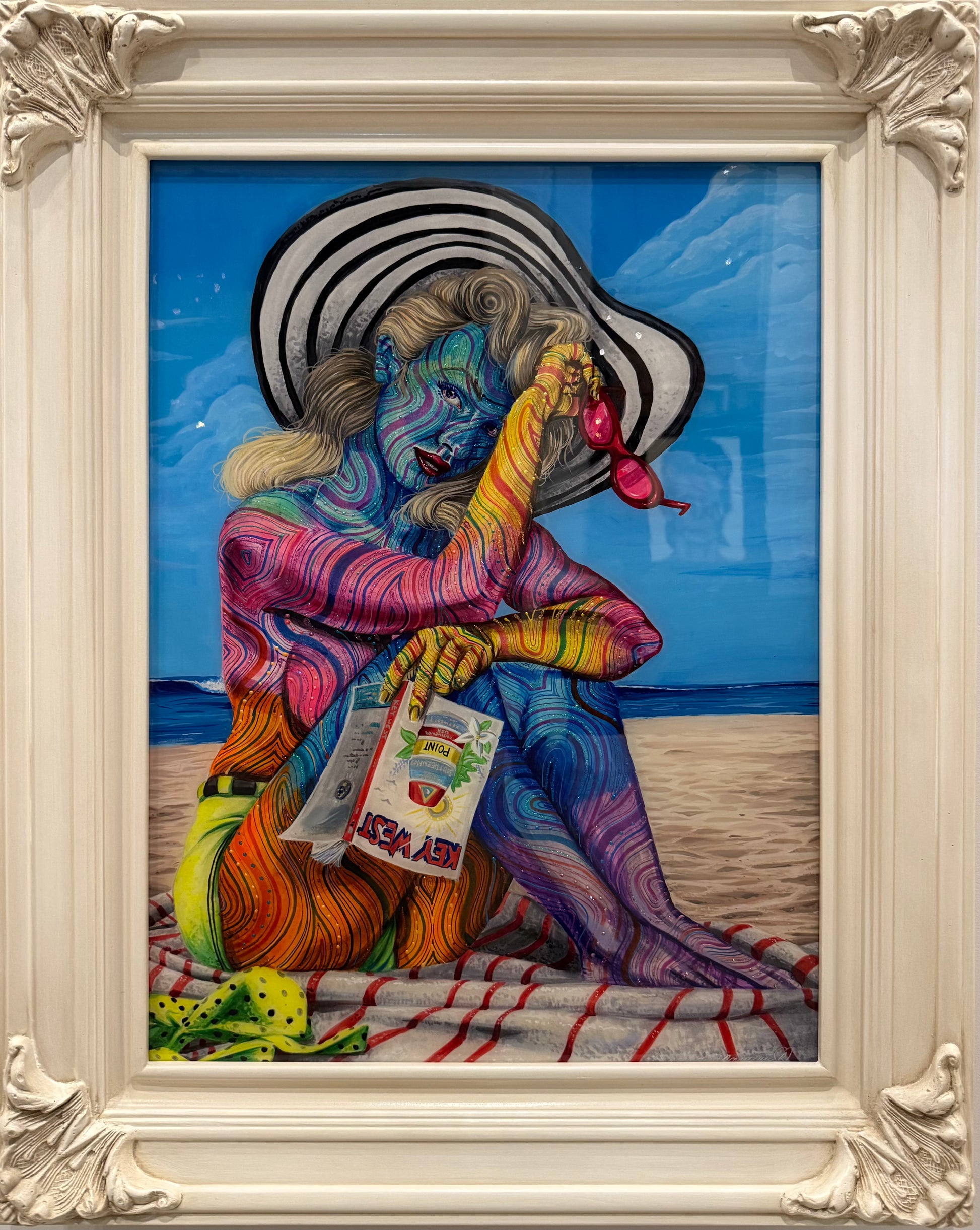 A framed colorful painting depicting a seated woman on a beach with a striped sun hat, blue sky, sand, and a red-and-white striped blanket. The ornate white frame surrounds the artwork.