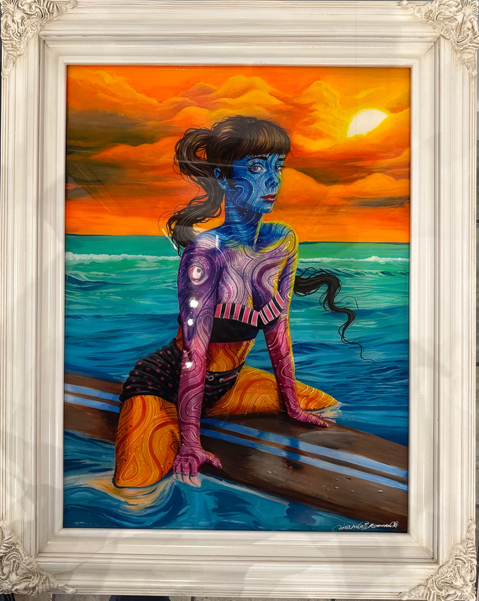 A framed surreal painting of a blue-skinned woman kneeling on a surfboard in the ocean under a bright orange sunset sky. The artwork is housed in an ornate white carved frame, shown hanging on a light wall.